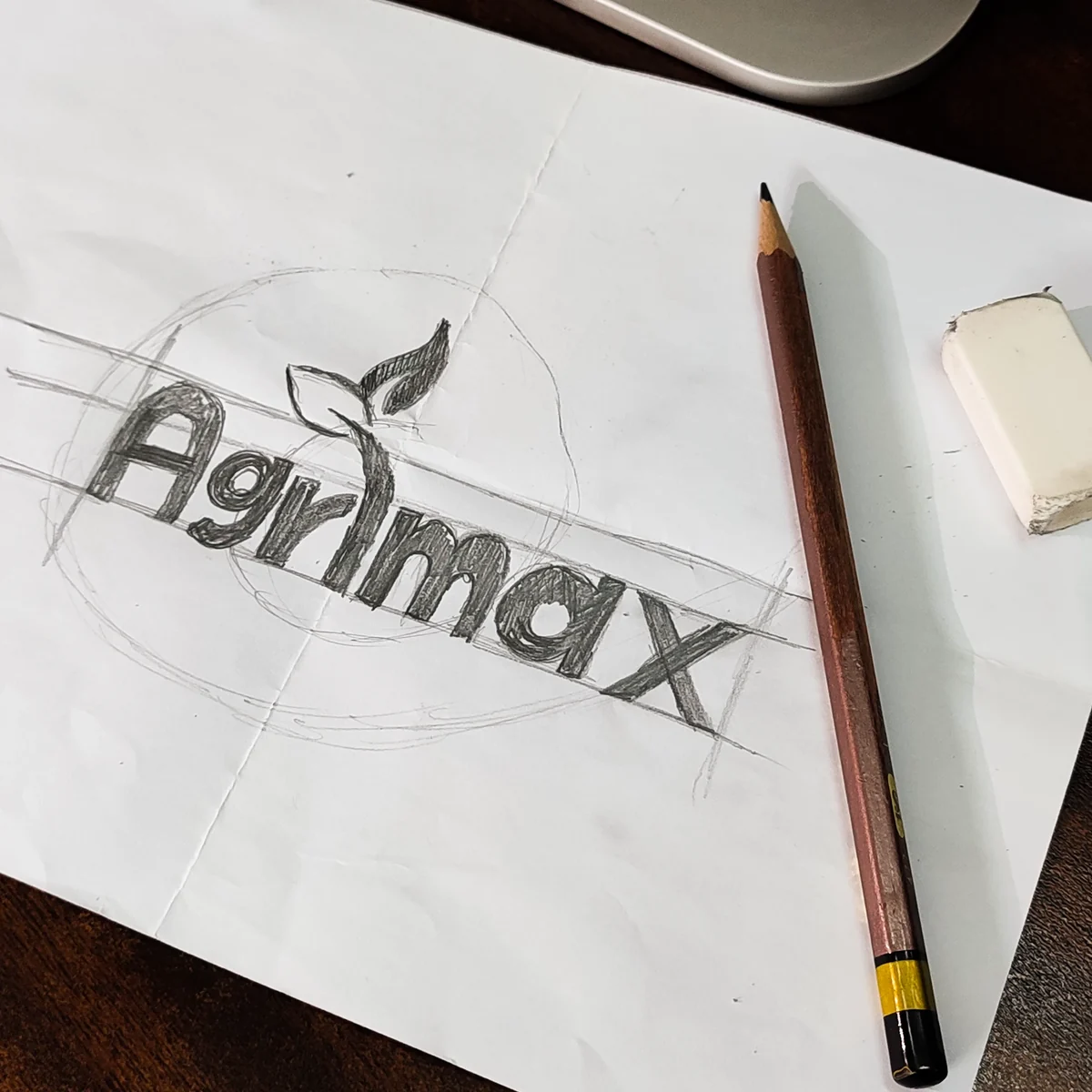 Pencil sketch of the Agrimax logo on paper with construction lines, a pencil and an eraser